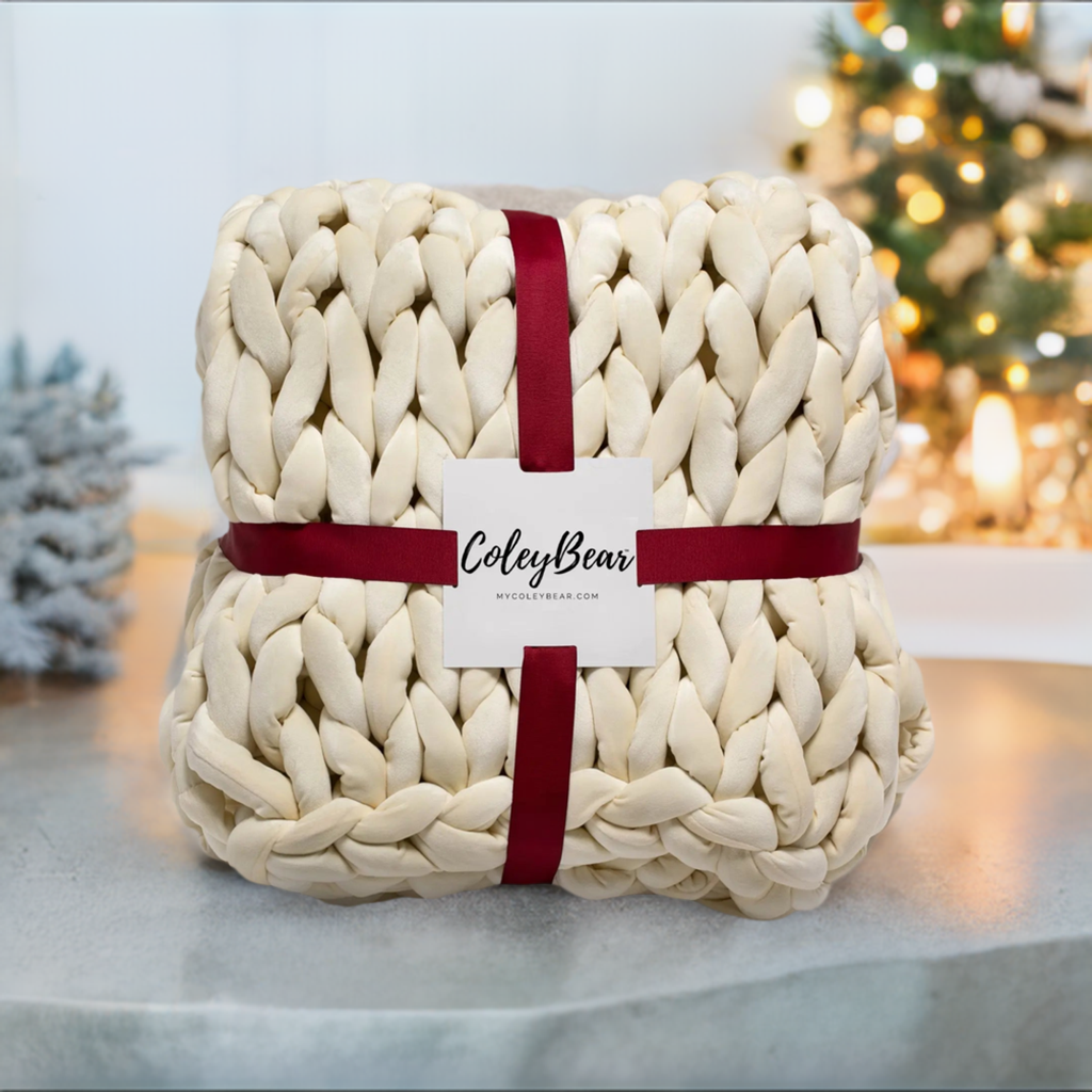 cream colored chunky knit weighted blanket wrapped in a burgundy ribbon and a ColeyBear card on the front. In the background there is a christmas tree.